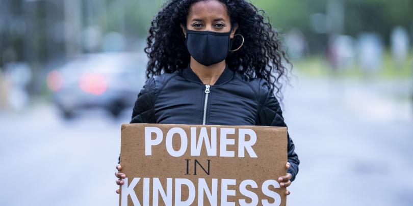 Woman in mask holding sign that says Power in Kindness