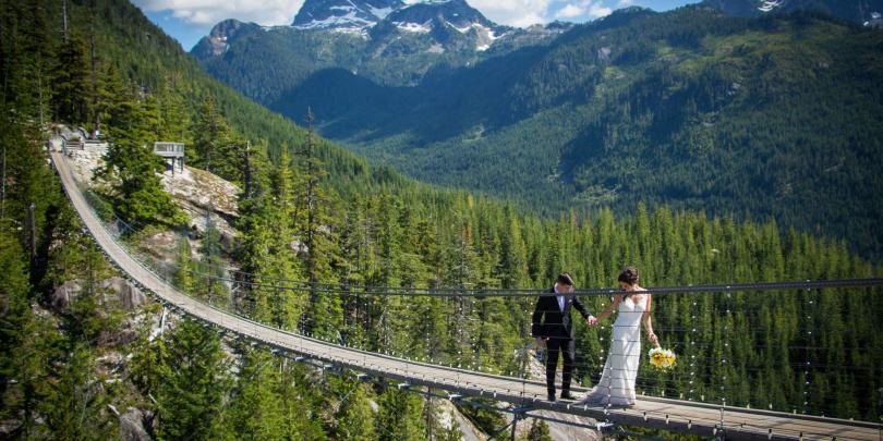 Josh Meyer in tuxedo, walks with his wife, in wedding dress across a suspension bridge in the mountains on their wedding day